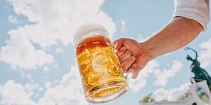 Männerarm reckt Maßkrug mit Bier in die Sonne, Hintergrund weiß-blauer Himmel