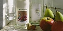 Gefülltes Likörglas mit Obstbrand, daneben zwei Flaschen fränkischer Obstler und frischen Birnen und Äpfeln